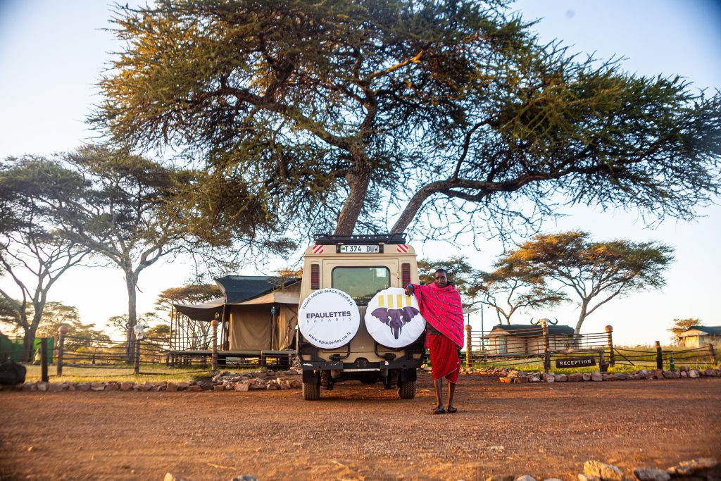 A proud Maasai warrior in traditional attire standing next to an Epaulettes Safaris car in the scenic savannah.